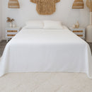 A neatly made bed is dressed in the Morgan and Reid Heavy-Weight Crisp Cotton Sheet Set, with pillows beside a white two-drawer nightstand holding vases with dried grasses. The gray wood floor shows part of a white rug.