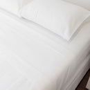 A neatly made bed is dressed in the Morgan and Reid Heavy-Weight Crisp Cotton Sheet Set, with pillows beside a white two-drawer nightstand holding vases with dried grasses. The gray wood floor shows part of a white rug.