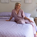 A neatly made bed features the Morgan and Reid Mosaic Lilac Coverlet Set with matching pillows, wooden nightstands, minimalist art above, and a vase of dried grass. The lightweight coverlet from Morgan and Reid is machine washable for easy care.