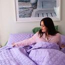 A neatly made bed features the Morgan and Reid Mosaic Lilac Coverlet Set with matching pillows, wooden nightstands, minimalist art above, and a vase of dried grass. The lightweight coverlet from Morgan and Reid is machine washable for easy care.