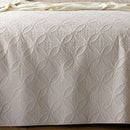 A neatly made bed with the Morgan and Reid Petal Oat Milk Coverlet Set, featuring a premium microfibre coverlet, two large pillows, a small white cushion, a beige upholstered headboard, bedside lamp, and potted plant on a wooden nightstand.