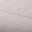 A neatly made bed with the Morgan and Reid Petal Oat Milk Coverlet Set, featuring a premium microfibre coverlet, two large pillows, a small white cushion, a beige upholstered headboard, bedside lamp, and potted plant on a wooden nightstand.