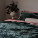 Bed with green velvet bedding and pink pillows, vase with plants on nightstand.