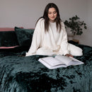 Woman in a white robe reading a book on a dark green bedspread.