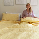 The Morgan and Reid Serene Butter Coverlet Set by Morgan and Reid, featuring a yellow coverlet and two matching pillows, brightens the room. A wooden nightstand with dried flowers and minimalist line art complete the cozy look.