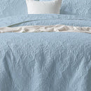 A neatly made bed features the Morgan and Reid Serene Corydalis Coverlet Set with embossed design and matching pillows, flanked by wooden nightstands with glass lamps. Two botanical prints hang above the headboard on a white paneled wall.