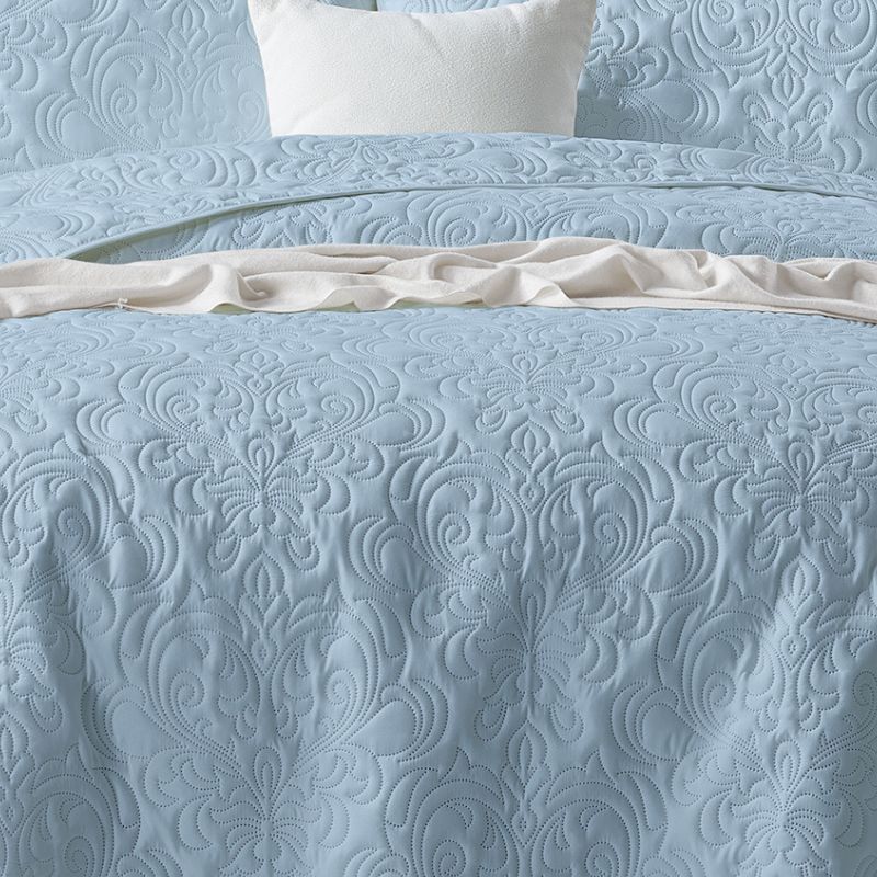 A neatly made bed features the Morgan and Reid Serene Corydalis Coverlet Set with embossed design and matching pillows, flanked by wooden nightstands with glass lamps. Two botanical prints hang above the headboard on a white paneled wall.