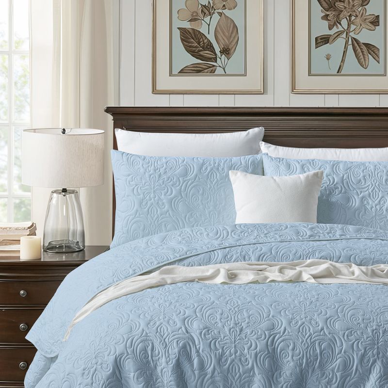 A neatly made bed features the Morgan and Reid Serene Corydalis Coverlet Set with embossed design and matching pillows, flanked by wooden nightstands with glass lamps. Two botanical prints hang above the headboard on a white paneled wall.