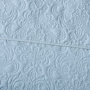 A neatly made bed features the Morgan and Reid Serene Corydalis Coverlet Set with embossed design and matching pillows, flanked by wooden nightstands with glass lamps. Two botanical prints hang above the headboard on a white paneled wall.