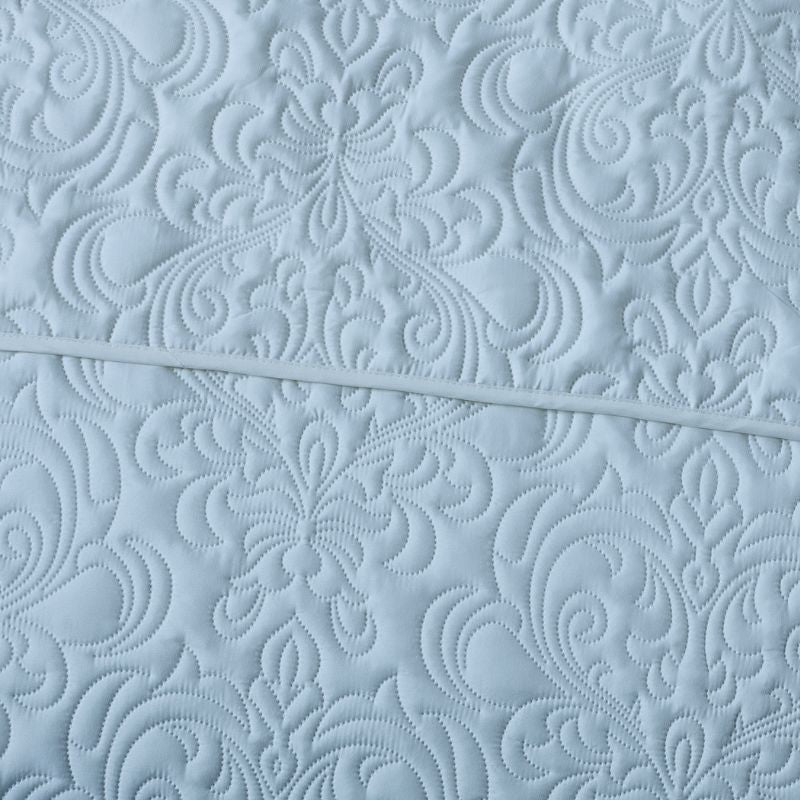 A neatly made bed features the Morgan and Reid Serene Corydalis Coverlet Set with embossed design and matching pillows, flanked by wooden nightstands with glass lamps. Two botanical prints hang above the headboard on a white paneled wall.