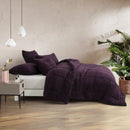 A neatly made bed with the Morgan and Reid Plum Snuggle Fleece Comforter Set and pillows sits on a light rug. Pendant lights hang above, with a side table, vase, and a large potted plant against a textured beige wall.