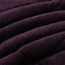 A neatly made bed with the Morgan and Reid Plum Snuggle Fleece Comforter Set and pillows sits on a light rug. Pendant lights hang above, with a side table, vase, and a large potted plant against a textured beige wall.