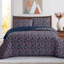 A neatly made bed with a dark floral comforter and matching pillows, the Morgan and Reid Sophia Coverlet Set in lightweight breathable fabric at the foot, a wooden headboard, geometric wall art, and a vase of flowers on the bedside table.