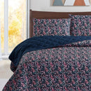 A neatly made bed with a dark floral comforter and matching pillows, the Morgan and Reid Sophia Coverlet Set in lightweight breathable fabric at the foot, a wooden headboard, geometric wall art, and a vase of flowers on the bedside table.