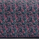 A neatly made bed with a dark floral comforter and matching pillows, the Morgan and Reid Sophia Coverlet Set in lightweight breathable fabric at the foot, a wooden headboard, geometric wall art, and a vase of flowers on the bedside table.