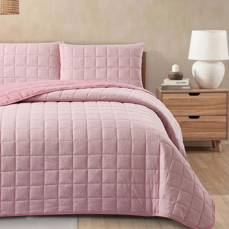 Pink quilted bedding set on a bed with a wooden nightstand and lamp in the background.