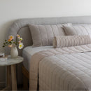 Neatly made bed with beige bedding and striped pillow, side table with flowers in a room.