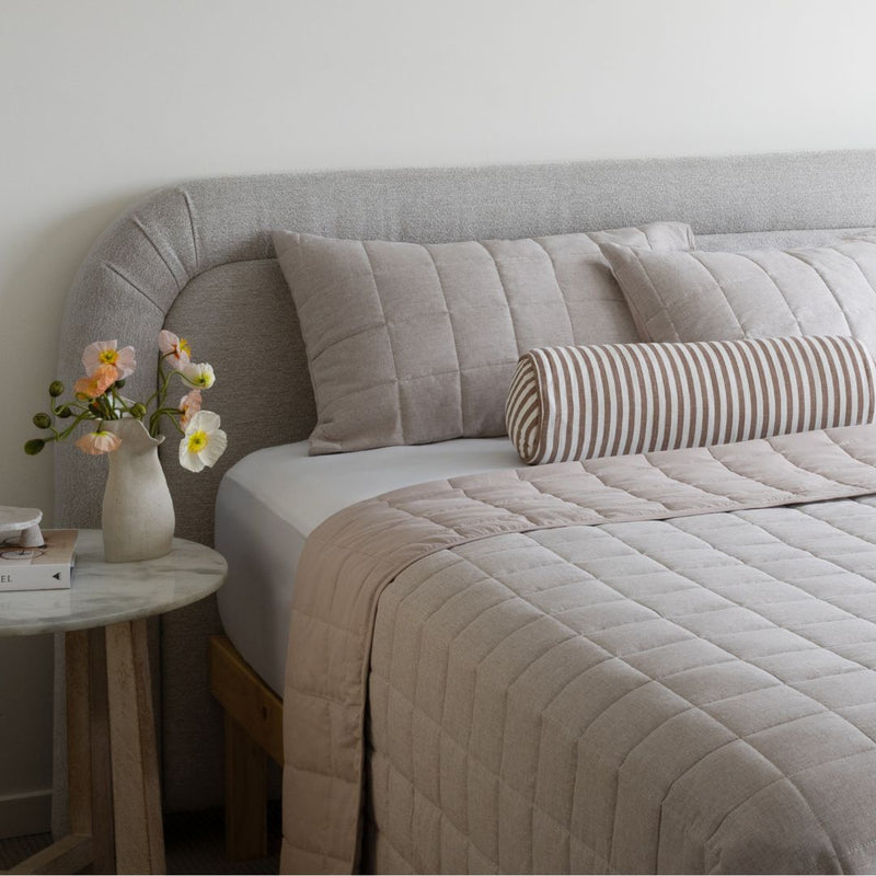 Neatly made bed with beige bedding and striped pillow, side table with flowers in a room.