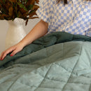 Person wearing a checkered shirt sitting on a bed with green bedding.
