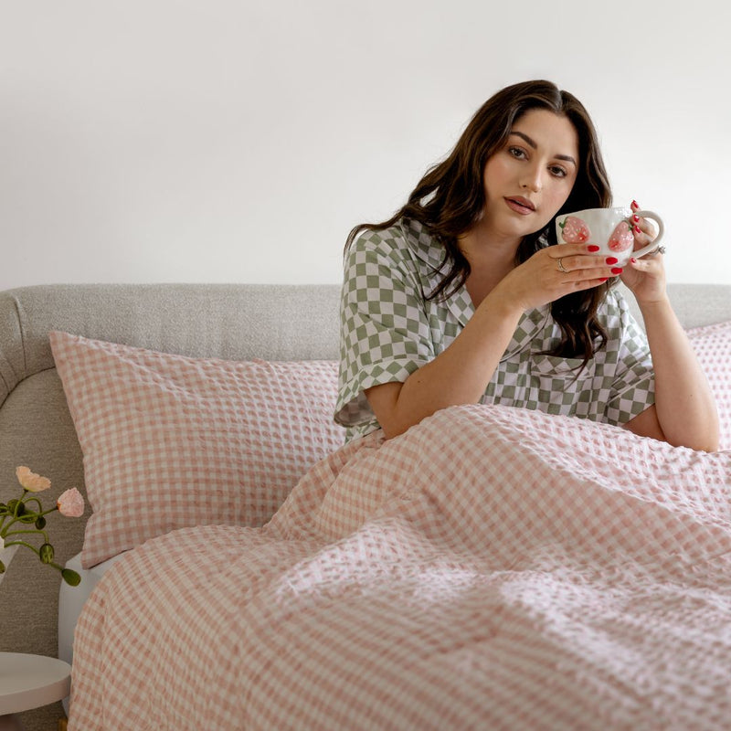 Woman in checkered pajamas holding a mug in bed with pink and white checkered bedding.