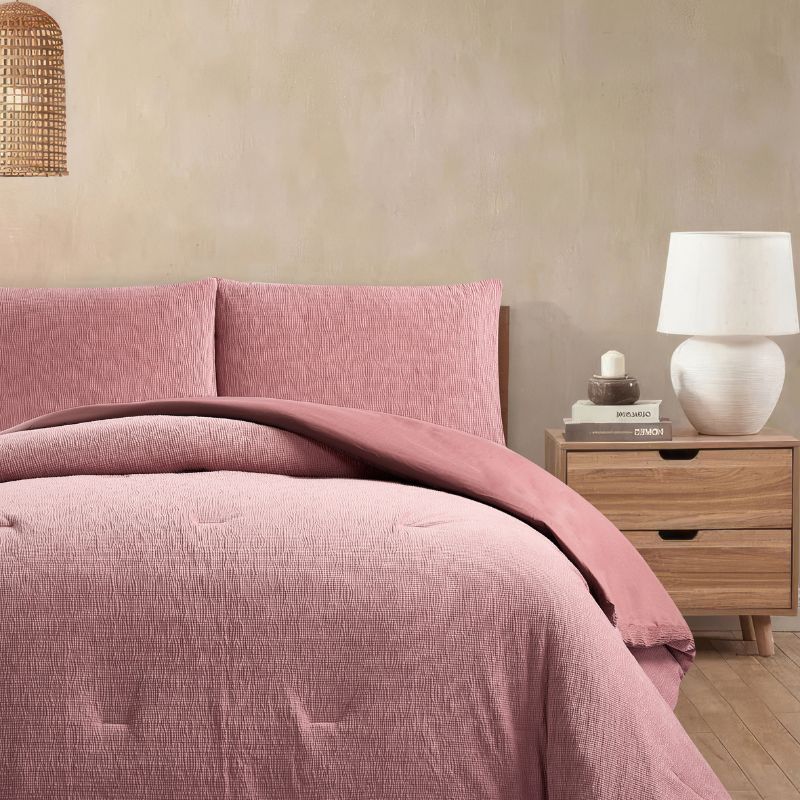 A modern bedroom features the Morgan and Reid Rose Stretch Woven Comforter Set on a neatly made bed with matching pillows, warm neutral walls, a woven pendant light, a wooden side table with a glass vase of white stems, and a ceramic lamp on a timber bedside table.