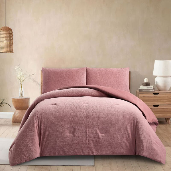 A modern bedroom features the Morgan and Reid Rose Stretch Woven Comforter Set on a neatly made bed with matching pillows, warm neutral walls, a woven pendant light, a wooden side table with a glass vase of white stems, and a ceramic lamp on a timber bedside table.