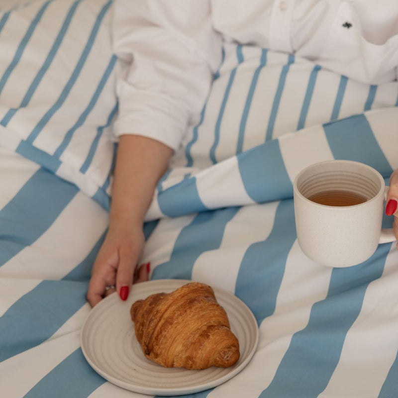 Person in pajamas holding a cup of tea and a croissant on a blue and white striped blanket.