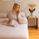 Woman wrapped in a blanket sitting on a bed next to a wooden nightstand with decorative elements.