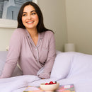 Woman in pink pajamas sitting on a bed with a bowl of fruit and a book.