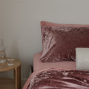 Bed with pink velvet bedding and pillows next to a wooden side table with a glass of water.