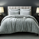 A neatly made bed features the Morgan and Reid Silver Velvet Crushed Snuggle Fleece Comforter Set with matching pillows, flanked by dark nightstands and modern lamps. A white cup and saucer sit on the comforter. The wall is dark gray.