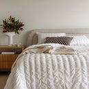 Neatly made bed with white bedding and a decorative pillow, nightstand with plant and books in a bedroom setting.