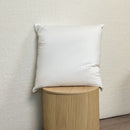 The Puradown 80% Duck Down 20% Duck Feather Cushion Insert by Puradown, in white, rests against a pale textured wall atop a grooved round wooden stool on a light wood floor.