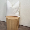 The Puradown 100% Duck Feather Cushion Insert by Puradown is displayed upright on a light wooden, ribbed cylindrical stool, set against a cream-colored textured wall and wooden floor.