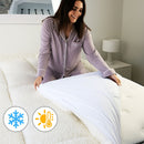 A modern bedroom features a light upholstered headboard, Sienna Living Luxury Hotel 1500GSM Reversible Mattress Topper, black bed base with wooden legs, a bedside table and lamp, with sunlight streaming through sheer curtains.