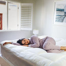 A modern bedroom features a light upholstered headboard, Sienna Living Luxury Hotel 1500GSM Reversible Mattress Topper, black bed base with wooden legs, a bedside table and lamp, with sunlight streaming through sheer curtains.