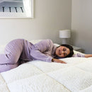 A modern bedroom features a light upholstered headboard, Sienna Living Luxury Hotel 1500GSM Reversible Mattress Topper, black bed base with wooden legs, a bedside table and lamp, with sunlight streaming through sheer curtains.