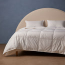 A neatly made bed features the Sienna Living Luxury 4 Seasons Goose Feather Down 2-in-1 Quilt by Sienna Living for year-round comfort. Two pillows rest against a curved beige headboard and blue wall above a light brown floor.