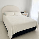 A minimalist bedroom features a neatly made bed with white bedding and two pillows, topped with the Sienna Living Summer Bamboo Cooling 2 in 1 Comforter Quilt. A lamp sits on a small nightstand, and sheer white curtains cover the window.