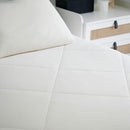 A minimalist bedroom features a neatly made bed with white bedding and two pillows, topped with the Sienna Living Summer Bamboo Cooling 2 in 1 Comforter Quilt. A lamp sits on a small nightstand, and sheer white curtains cover the window.