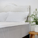 A neatly made bed with white sheets and pillows, a wicker headboard, and the Sienna Living Bamboo Mattress Protector. A small wooden table beside the bed holds a white vase filled with green leafy branches.