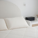 A neatly made bed with white pillows and Sienna Living Cooling 2 in 1 Pillowcase Pair sits beside a white nightstand with a black tissue box and beige bowl, all set against a minimal white wall for a refreshing sleep surface.