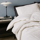 A neatly made bed with Sienna Living All Seasons 50% Duck Down 50% Duck Feather Quilt, white bedding and pillows sits by a black nightstand with a white lamp and two stacked books, set against a blue wall.