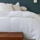 A neatly made bed features the Sienna Living All Seasons Hungarian Goose Down Quilt and white pillows against a dark blue paneled wall. A wooden bench at the foot of the bed adds all-season versatility to this clean, modern look.