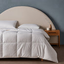 A bed dressed in white striped bedding and pillows showcases the Sienna Living Ultra Warm Winter Hungarian Goose Down Quilt, beside a wooden nightstand with a white mug and vase, set against a blue-gray wall and arched beige headboard.