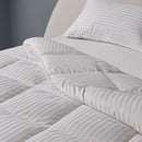 A bed dressed in white striped bedding and pillows showcases the Sienna Living Ultra Warm Winter Hungarian Goose Down Quilt, beside a wooden nightstand with a white mug and vase, set against a blue-gray wall and arched beige headboard.