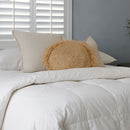 A modern bedroom with a neatly made bed featuring the Sienna Living Winter Hungarian Goose Down Quilt. Behind the bed is a blue accent wall, round mirror, simple decor, and wall sconces. A wooden bench with books sits at the foot of the bed.