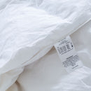 A modern bedroom with a neatly made bed featuring the Sienna Living Winter Hungarian Goose Down Quilt. Behind the bed is a blue accent wall, round mirror, simple decor, and wall sconces. A wooden bench with books sits at the foot of the bed.