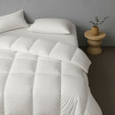 A bright, minimalist bedroom with a white bed topped by the Sienna Living Winter 80% Duck Down 20% Duck Feather Quilt, beside a wooden nightstand with an open book and shelf displaying wooden arches and a dark vase with a branch.