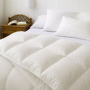 A neatly made bed features the Sienna Living Winter 80% Goose Down 20% Goose Feather Quilt from Sienna Living, paired with two pillows, set against a plain light blue wall on a light-colored carpeted floor.